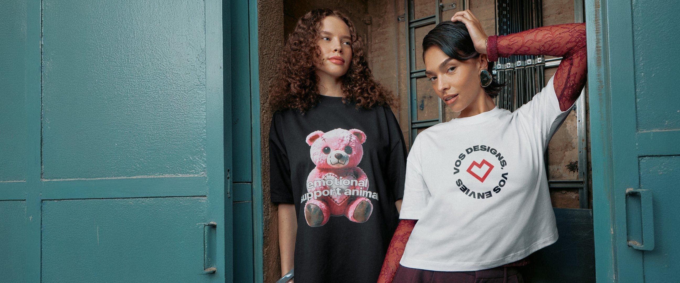 Deux femmes posent dans un espace industriel ; l’une porte un t-shirt noir avec un ours rose, l’autre un t-shirt blanc avec logo circulaire.