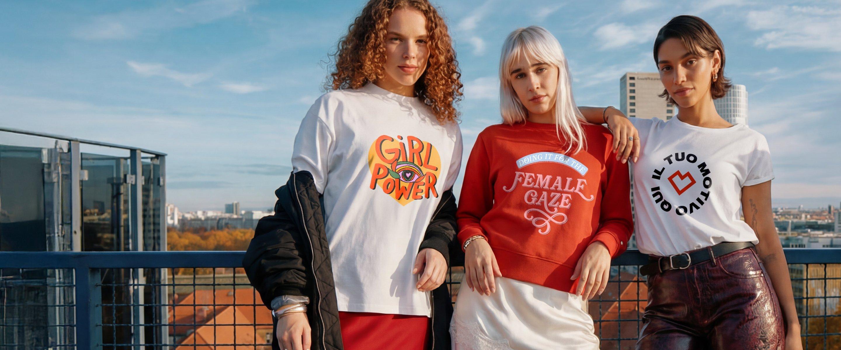 Tre donne si trovano su una terrazza panoramica con vista sulla città e indossano magliette con slogan femministi come “Girl Power”, “Female Gaze”, oltre al testo personalizzabile “Il tuo motivo qui”.