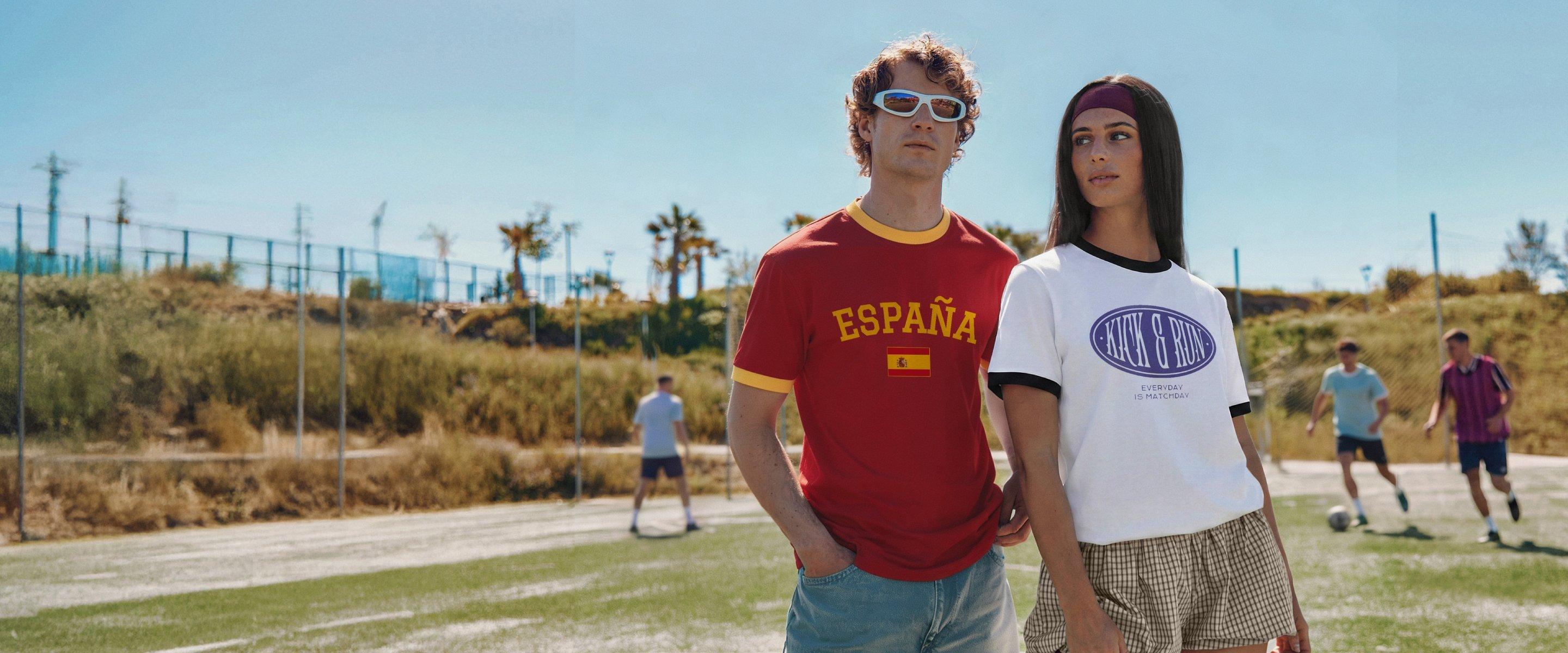 Two people stand on a football pitch wearing printed retro shirts with football designs while a match is played in the background.