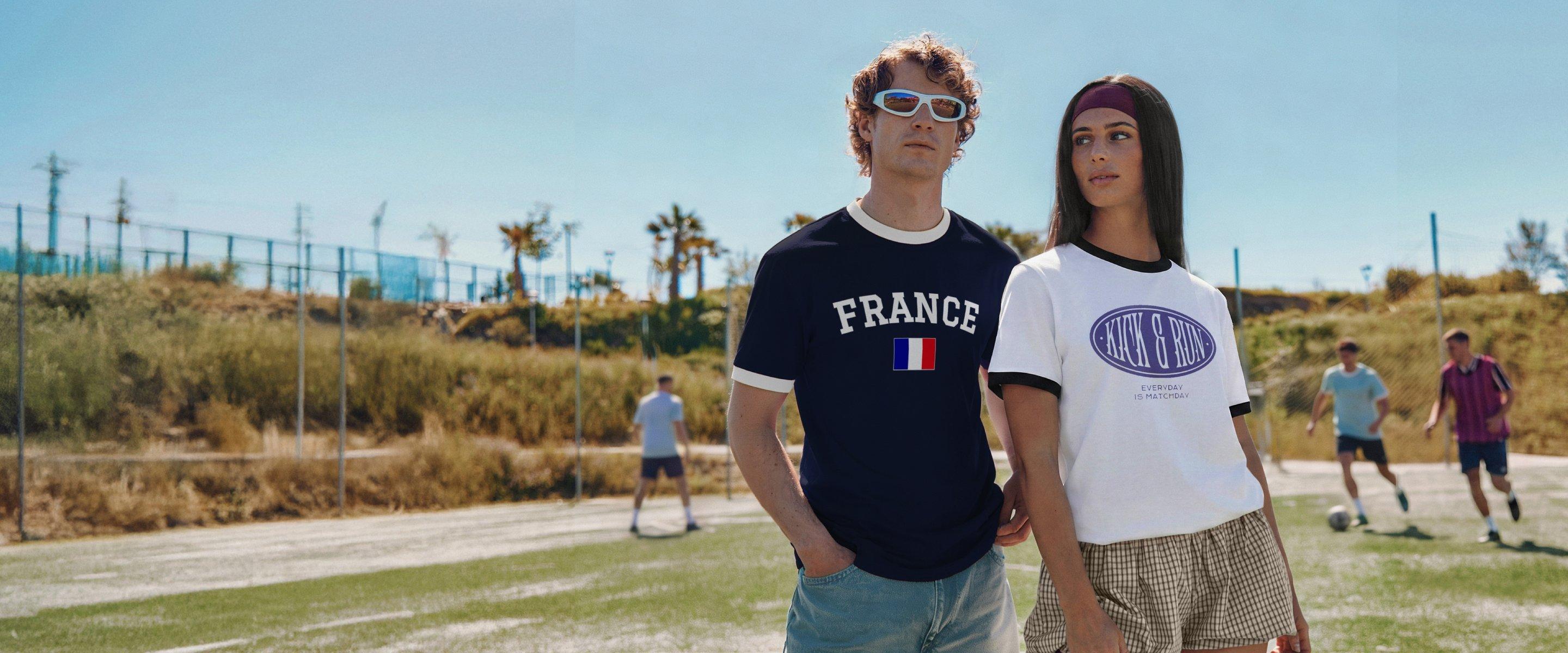 Deux personnes se tiennent sur un terrain de football avec des t-shirts rétro imprimés sur le thème du football pendant qu’un match se joue en arrière-plan.