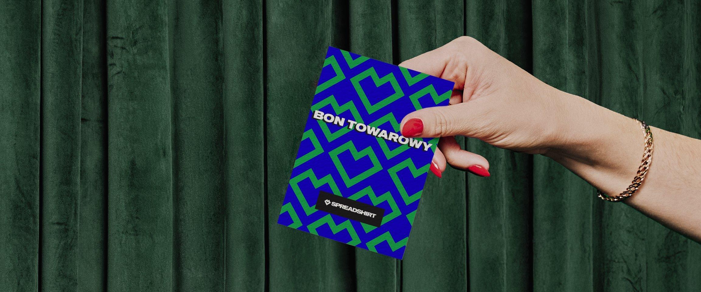 Niebieska karta z zielonym wzorem serc i białym napisem „Bon Towarowy” oraz „Spreadshirt” jest trzymana w dłoni na tle zielonego tła.