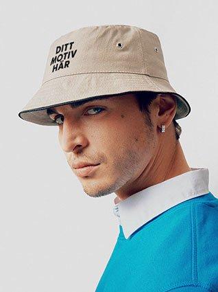 Man med beige fiskehatt, svart tryck "Ditt motiv här", porträtt fotograferat från sidan.