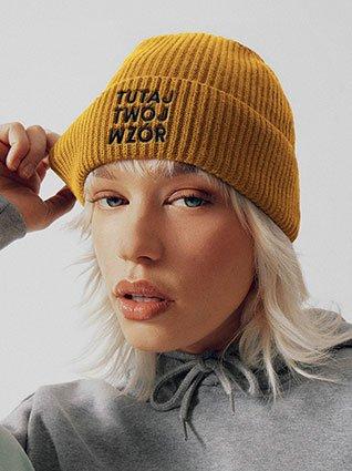 Kobieta w żółtej czapce z dzianiny, z czarnym haftem „Twój wzór tutaj” z przodu.