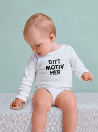 Baby i hvit langermet body med svart påtrykk «Ditt motiv her» sitter og ser ned.