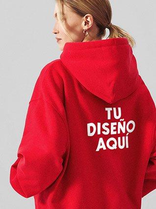 Persona de cabello recogido usando sudadera roja con capucha y el texto "TU DISEÑO AQUÍ" en la espalda.