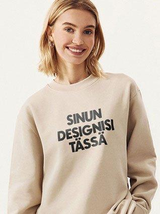 Nainen hymyilee beigen värisessä puserossa, jossa musta painatus ”Sinun designisi täällä”.