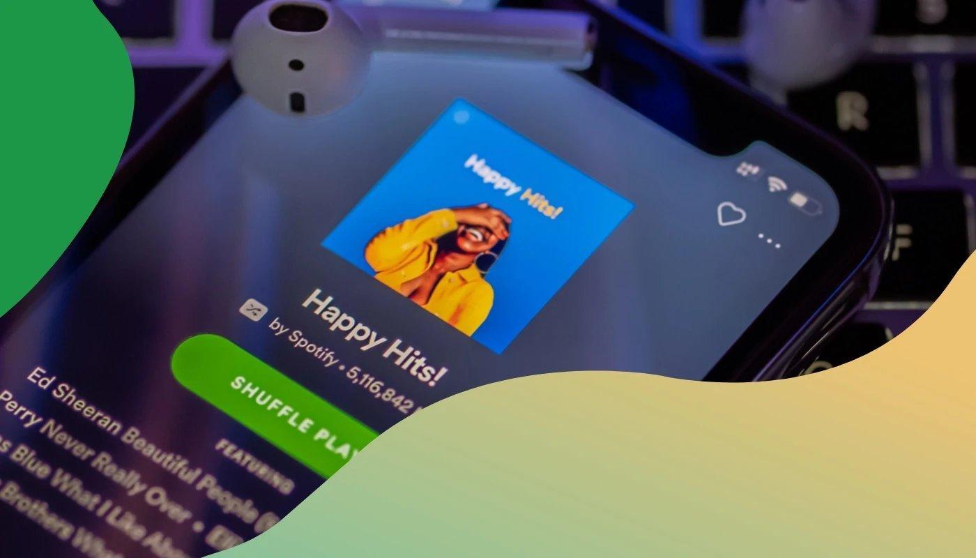 Smartphone mit geöffneter Spotify-App zeigt die Playlist „Happy Hits!“ mit einem lachenden Model auf dem Cover, darüber liegt ein kabelloser Kopfhörer.
