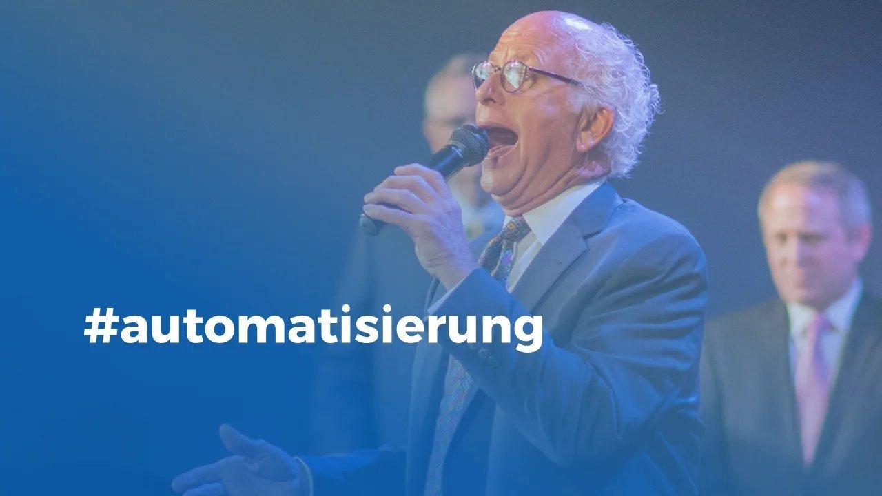 Drei alte weiße Männer in Anzug stehen auf einer Bühne wobei ein Mann eine Rede hält und in ein Mikrofon spricht.