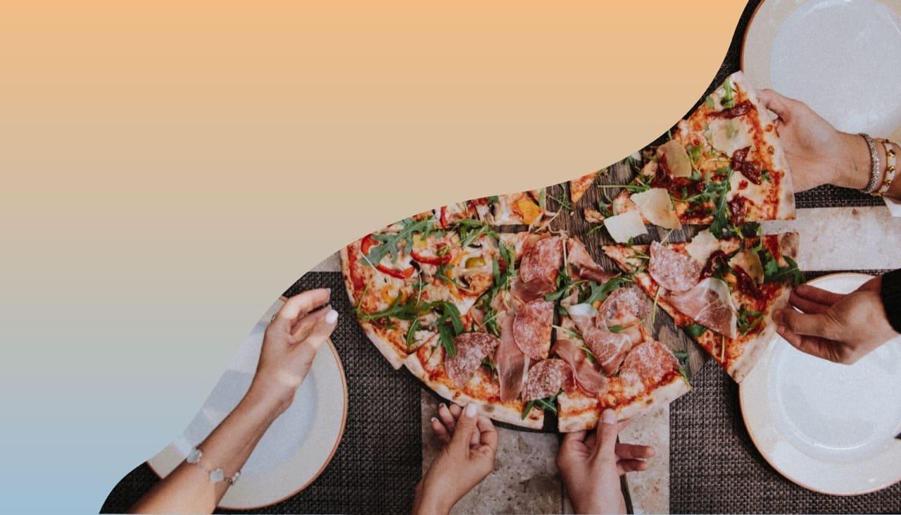 Draufsicht auf einen Tisch mit großer geschnittener Pizza in der Mitte und vieler Hände, die sich jeweils ein Stück der Pizza greifen.