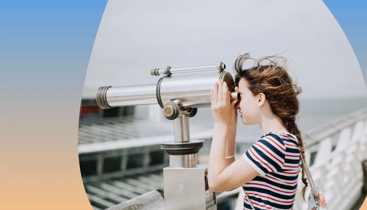 Junges Mädchen, das auf einem Steg durch ein Einwurf-Fernglas in die Weite schaut.