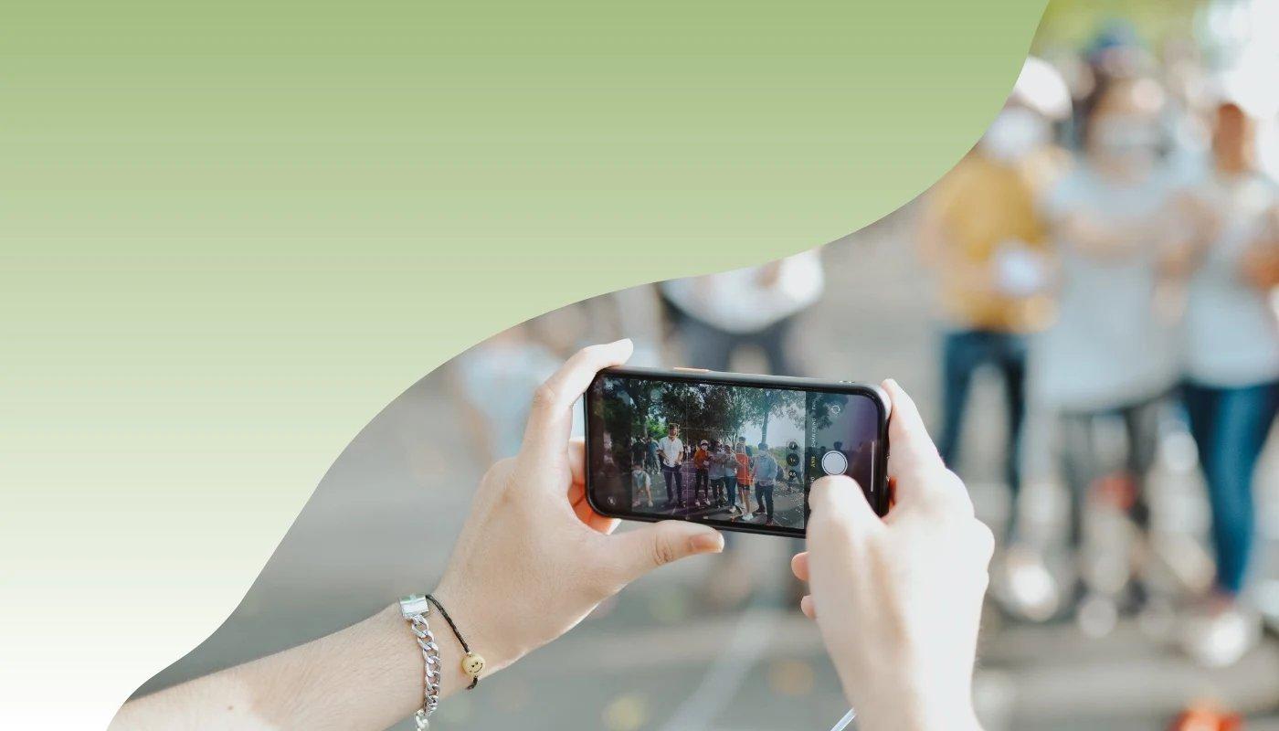 Nahaufnahme einer Person, die mit dem Smartphone eine Gruppe Menschen filmt, unscharfer Hintergrund mit Outdoor-Szene.