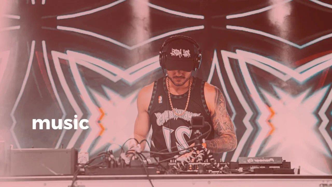 DJ auf einer Bühne steht hinter dem DJ pult und bedient die Regler während im Hintergrund visuelle Effekte und Lichter die Bühne ausfüllen.