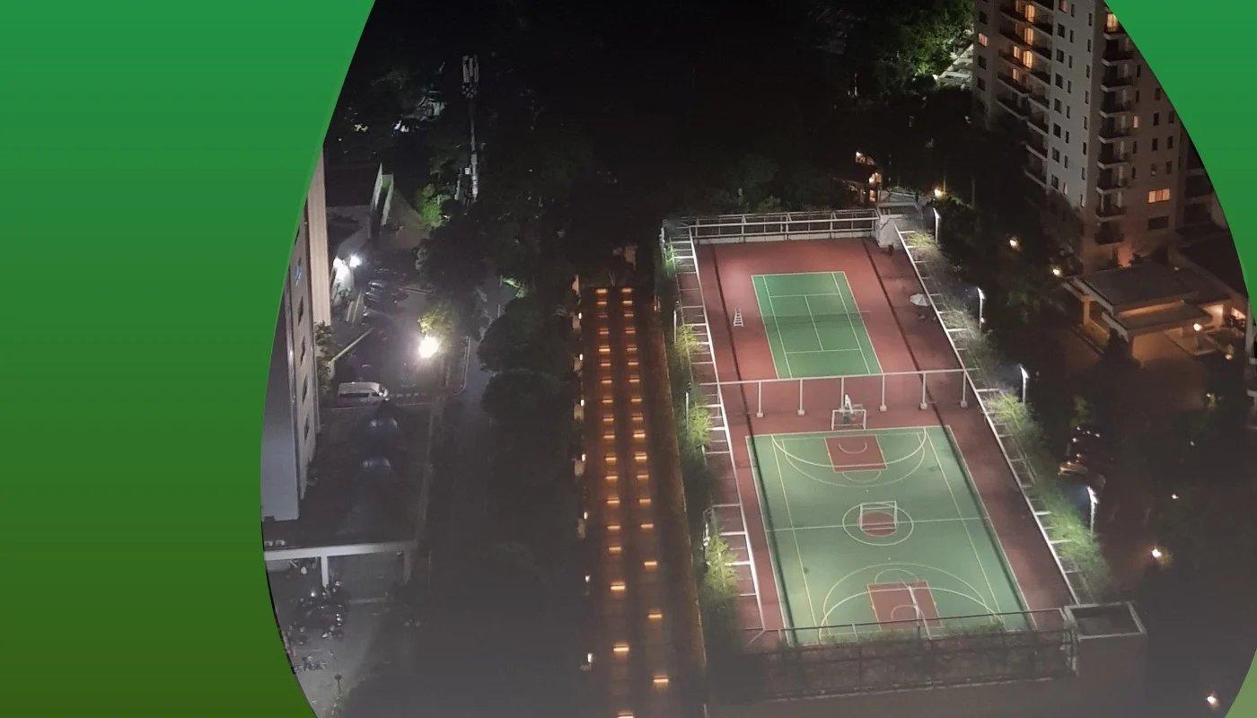 Sportplatz mit Basketball- und Tennisfeldern bei Nacht, von oben fotografiert, beleuchtet zwischen Wohngebäuden.