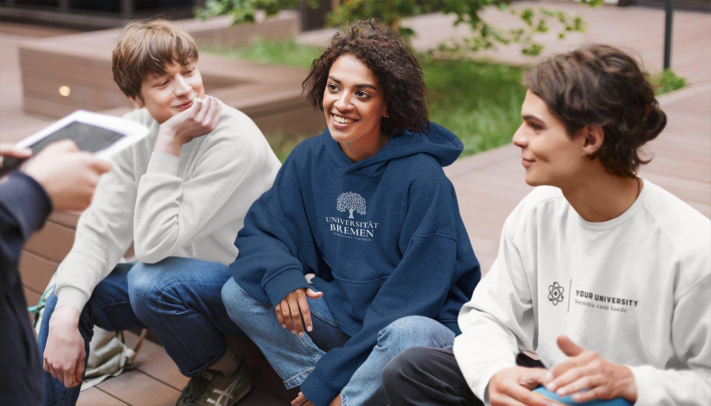 Drei junge erwachsene sitzen mit passendem Merch ihrer Hochschule auf dem Campusgelände.