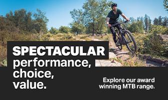 A man wearing a helmet and knee pads riding a mountain bike down a rocky trail in a woodland area.